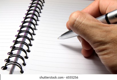 A Hand Holding A Pen Above A Blank Page Of A Spiral Notebook.