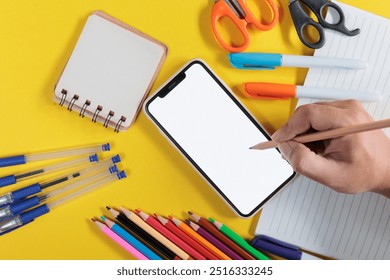 Hand holding pencil on smartphone screen and consists of notebook, magic pen, scissors placed on yellow surface. Education concept - Powered by Shutterstock