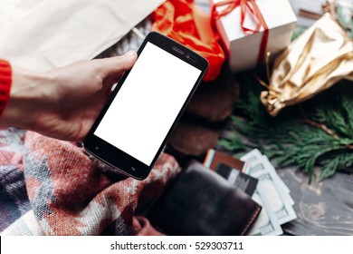 Hand Holding Phone With Empty Screen . Seasonal Sale. Christmas Shopping Concept. Rustic Background With Wallet And Bags Of Presents