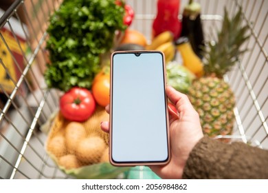 Hand Holding Phone With White Screen Copy Space In Grocery Shopping Cart With Fruits And Vegetables Healthy Food