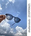 hand holding photochromic minus glasses against the background of a clear blue sky with scattered white clouds.