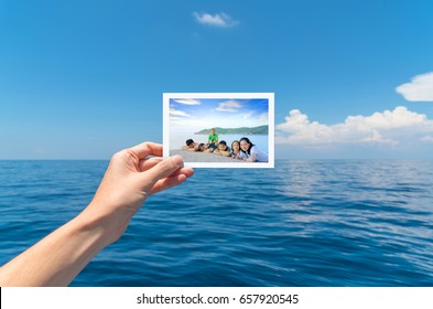 Hand Holding A Picture Of Happy Big Asian Family Tourist On Vacation With Beautiful White Clouds On Blue Sky Over Calm Sea In Background.