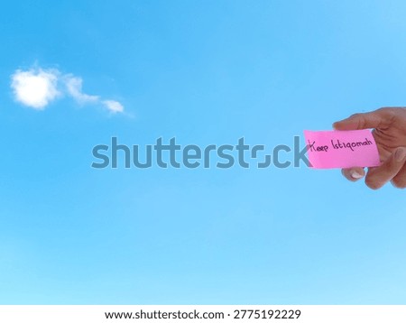 A hand holding a pink note with 'Keep Istiqomah' text against a clear blue sky. Concept of consistency, steadfastness in faith, and persistence in achieving goals