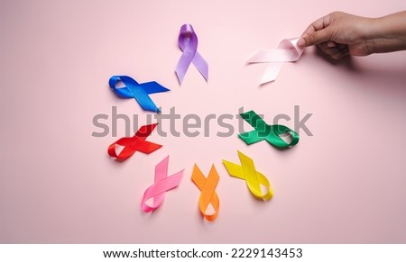 Hand holding pink ribbon which is among colorful ribbons on pink background, cancer awareness, World cancer day, National cancer survivor day, world autism, supported living and illness.