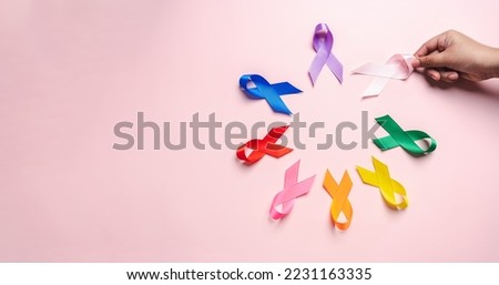 Hand holding pink ribbon which is among colorful ribbons on pink background, cancer awareness, World cancer day, National cancer survivor day, world autism, supported living and illness.