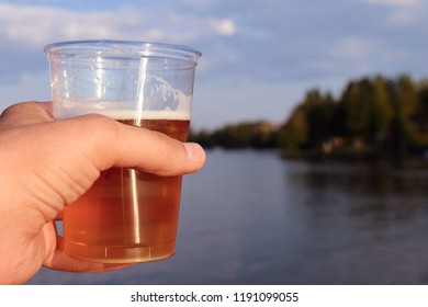 Hand Holding A Plastic Beer Mug. Summer Lake On Background.