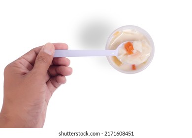 Hand holding plastic spoon scooping fresh milk tofu with fruit salad topping in plastic cup to taste isolated on white background. Top view