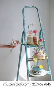 Hand Holding A Plate With A Cake, Next To It Incredible Beautiful Cakes With A Cut Decorated With Fresh Flowers On A Ladder After Repair