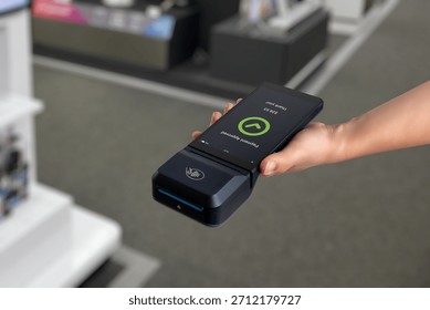 Hand holding POS terminal in electronics store, showing payment approved screen during digital checkout and cashless transaction process