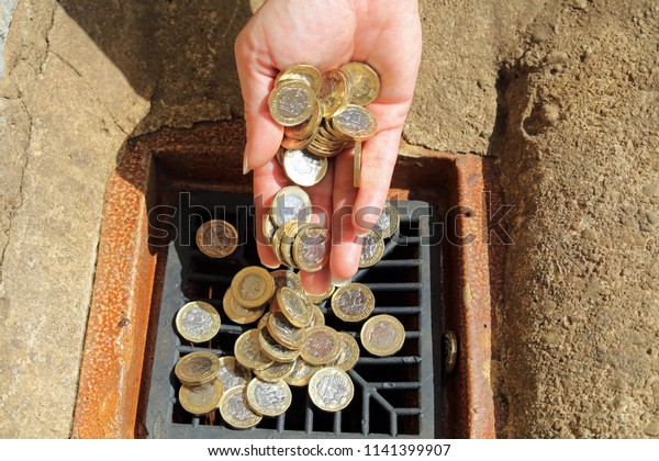 Hand Holding\
Pound Coins And Tipping Them Away. Pouring Money Down The Drain,\
Common Phrase For Wasting\
Money.