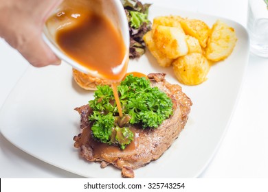 Hand Holding And Pouring Gravy Sauce On Ribeye Steak With Fried Potato And Bread