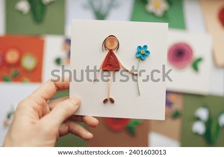 Hand holding quilling card with stick figure girl giving flower. woman making greeting cards. Hand made of paper quilling technique. Handicraft at home. Hobby, home office.