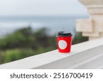 A hand holding a red coffee cup with a white circle design, set against a blurred outdoor background. The cup is positioned on a white ledge, suggesting a casual moment of enjoying a beverage.