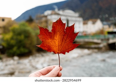 Hand holding a red maple leaf, picturesque Canadian landscape with a river and mountains.