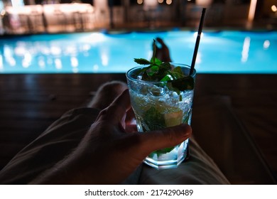 Hand Holding A Refreshing, Nicely Decorated Cocktail Next To The Illuminated Swimming Pool Of The Hotel At Night Time. Bar In The Blurry Background. Relaxing Summer Vacation Scene.