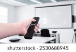 Hand holding a remote control, operating a projector in a conference room or classroom. Concept of presentation control, business meeting, education, technology