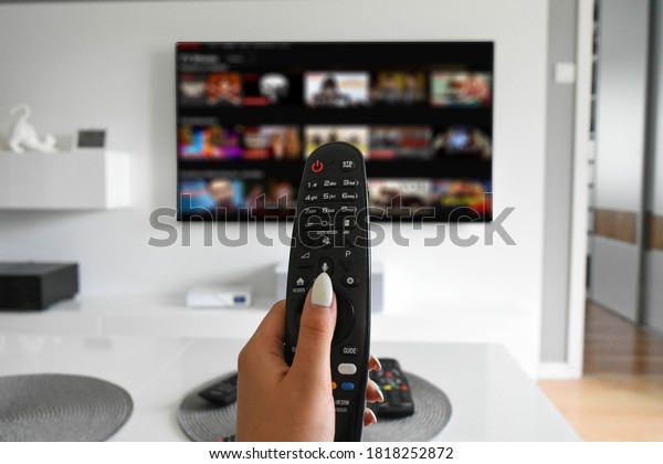 Hand holding a\
remote controller with a streaming website on TV in the blurred\
background, selective\
focus.
