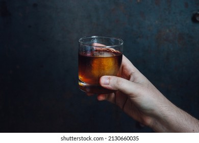 Hand Holding Rocks Glass Of Whiskey With Ice Against Dark Blue Metal Background. Copy Space.