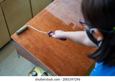 Hand holding roller and applying polyurethane on the wooden board by a young girl.
