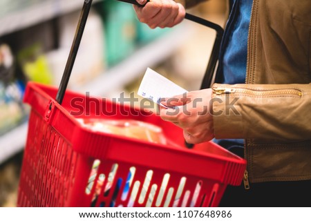 Hand holding shopping list and basket in grocery store aisle. Woman reading paper, shelf in the background. Lady buying groceries in supermarket. Consumer in hypermarket purchase cheap food.