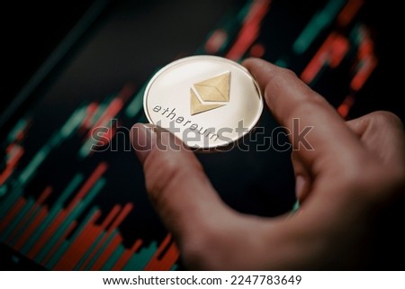 Hand holding a silver Ethereum (ETH) cryptocurrency coin with candle stick graph chart and digital background.