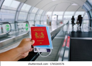 Hand holding Singapore passport, boarding pass and face mask, in airport. Person on travelator / travolator, Travel concept. Reopening; coronavirus covid-19; travel restrictions.
