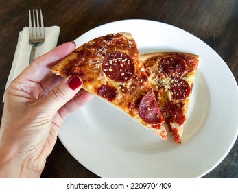 Woman’s Hand Holding A Slice Of Pepperoni Pizza Above And A Thin Crust Pizza Slice On A White Dinner Plate Below. 