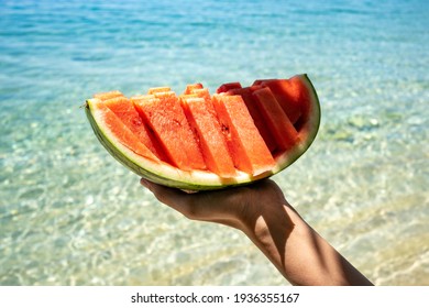 Hand holding slice of watermelon on the beach. Sea and blue sky background. Summer mood. 2021