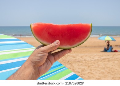 A Hand Holding A Slice Of Watermelon, On The Beach. Concept Of The Natural Way To Refresh Yourself..