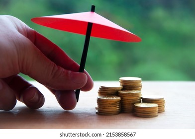 Hand holding small red umbrella over pile of gold coins on wooden table. Close up of stack of gold coins with hand holding the umbrella for protection. Financial safety and investment concept.