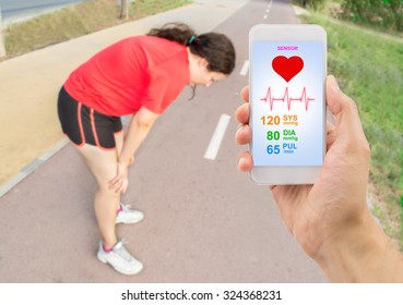 Hand Holding The Smart Phone With A Healthy App And In Background A Woman Gasping For Air With Her Hands On Her Knees After A Run