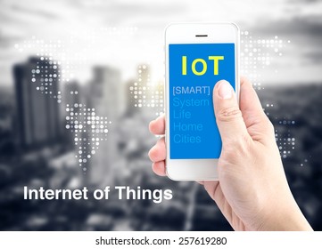 Hand Holding Smart Phone With Internet Of Things (IoT) Word With Dot World Map And City Blur Background, Digital Marketing Concept.