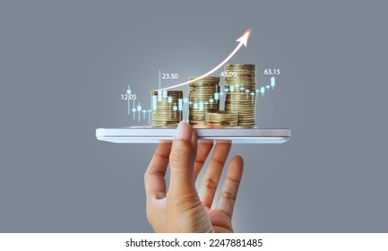 Hand holding smart phone and stack of coins over stock market screen and financial graph in background. Digital marketing hand holding smart phone with graphic icon - Powered by Shutterstock