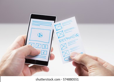 Hand holding smart phone, user interface designer checking wireframe sketch for mobile app. Prototype sketch of mobile responsive website