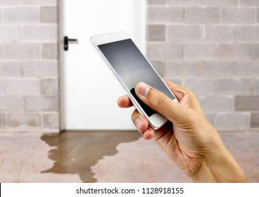 A hand holding up a smart phone with water damage in basement caused by sewer backflow due to clogged sanitary drain background 