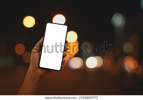 Hand holding smartphone with blank screen at\
night on bokeh city street\
background