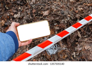 Hand Holding A Smartphone With Blank White Screen Against The Background Of Fallen Leaves And Red White Striped Warning Tape. Crime Scene Investigation
