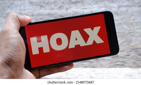Hand holding smartphone with letter HOAX on red background. Propaganda, disinformation and hoax concept.