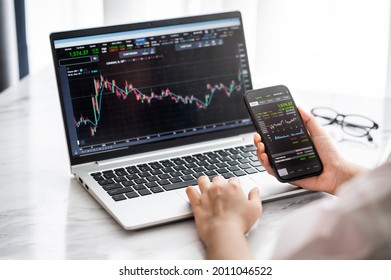 Hand holding smartphone with stock market data and using laptop display graph and chart for analyze and check before trading stocks online