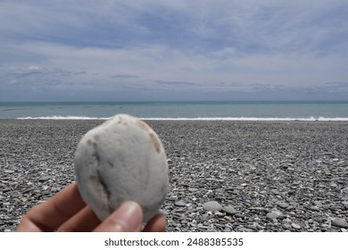 Hand holding a smooth white stone in focus with a pebbled beach and calm ocean in the background under a vast cloudy sky. - Powered by Shutterstock