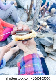 Hand Holding A Smore Made With An Oreo. Rocks And Camp Fire In Background With People Around It. 