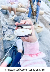 Hand Holding A Smore Made With An Oreo. Rocks And Camp Fire In Background With People Around It. 