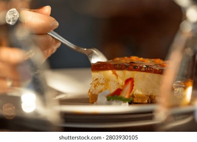 Hand holding a spoon, taking a bite from a delicious slice of passion fruit cheesecake garnished with strawberry and mint, served on a white plate in a restaurant setting