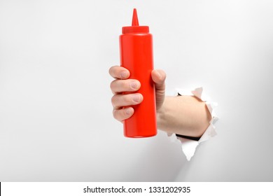 Hand Holding A Squeeze Bottle Of Ketchup Through Torn White Paper. Sauce, Condiment To Add Tomato Taste To Food.