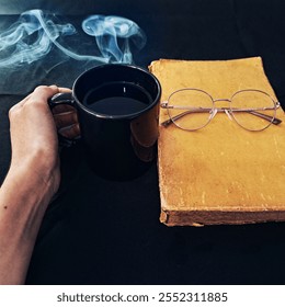 A hand holding a steaming coffee cup, with a vintage book and glasses on a dark background. The warm tones create a cozy and relaxing atmosphere - Powered by Shutterstock