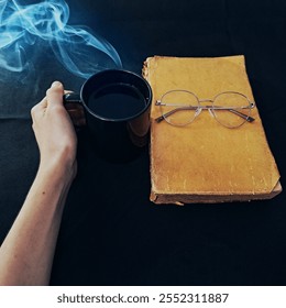 A hand holding a steaming coffee cup, with a vintage book and glasses on a dark background. The warm tones create a cozy and relaxing atmosphere - Powered by Shutterstock