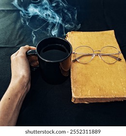 A hand holding a steaming coffee cup, with a vintage book and glasses on a dark background. The warm tones create a cozy and relaxing atmosphere - Powered by Shutterstock