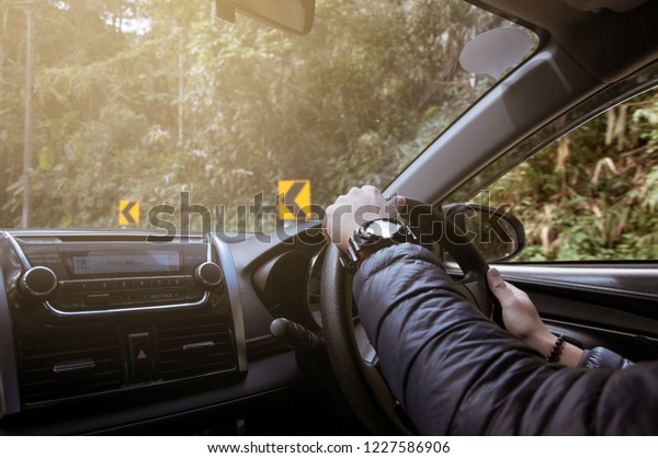 Men’s hand holding steering wheel of car while\
driving on the mountain\
road