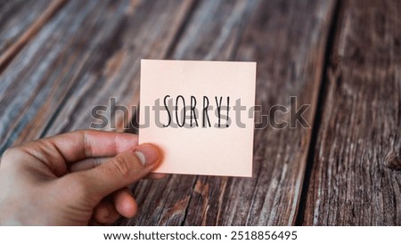 Hand holding sticky note that says 'SORRY!' against a rustic wooden background.