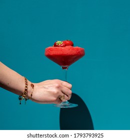 A hand holding a strawberry dairquiri cocktail in front off a blue background